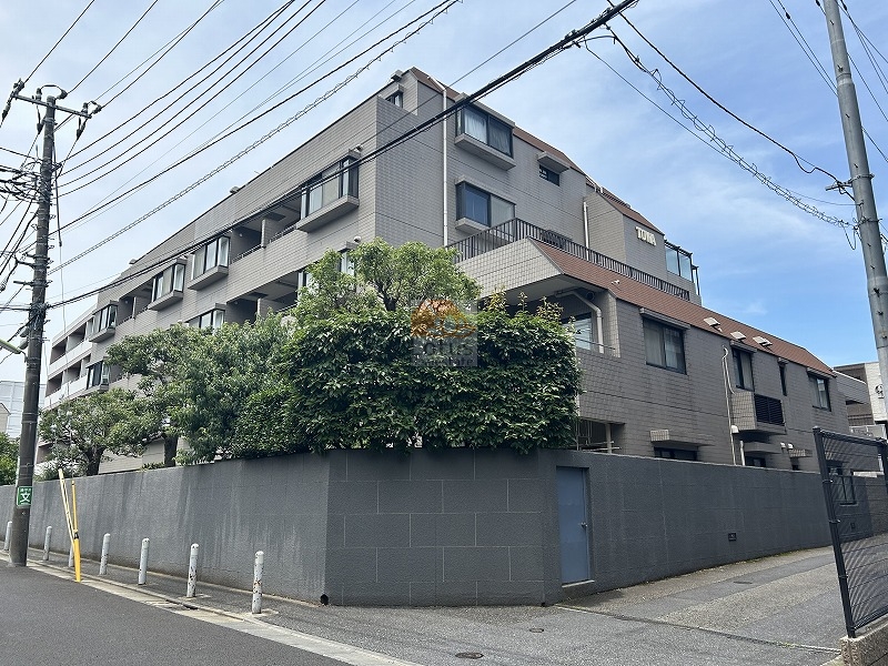 藤和上北沢ホームズの外観です。角を曲がると、グレーのタイル外壁の住棟が道路沿いに続き、段差のある勾配屋根が連なって見えます。周囲は高さのある塀と生垣で囲われ、電柱と配線が交差する街路が前面に伸びています。藤和上北沢ホームズは、昭和62年築、5階建てのマンションです。
