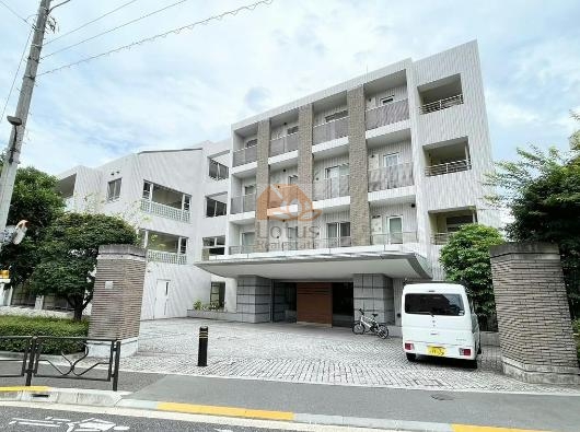 サンウッド文京開運坂上は白系の外壁に縦ラインの意匠を取り入れ、落ち着いた雰囲気を形成しています。エントランス前には大きな庇が設けられ、車寄せスペースと自転車が並ぶスペースが確認できます。道路に面した部分には植栽や門柱が整備され、住宅街の景観に調和した外観となっています。サンウッド文京開運坂上は、平成14年築、10階建てのマンションです。
