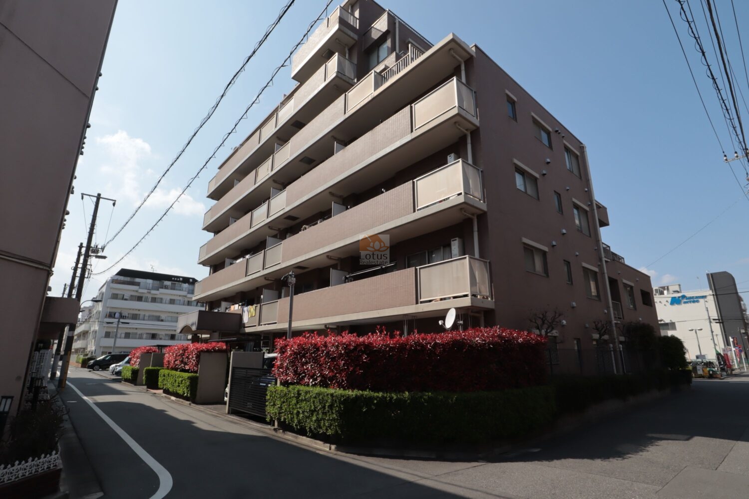 茶色を基調としたタイル張りの外観は、周囲の景観と調和しつつ、重厚感と安定感を感じさせます。手入れの行き届いた生け垣と、鮮やかな赤色が目を引く植栽が建物の足元を美しく彩り、住民を迎えます。南側からのアングルからは、各戸に設けられたバルコニーが陽光をたっぷりと取り込む様子が伺え、明るく快適な生活空間を予感させます。通りに面していながらも、植栽がプライバシーを適度に守る役割を果たしており、落ち着いた暮らしを実現します。都市の利便性を享受しながら、安らぎある住環境を求める方にふさわしいレジデンスです。