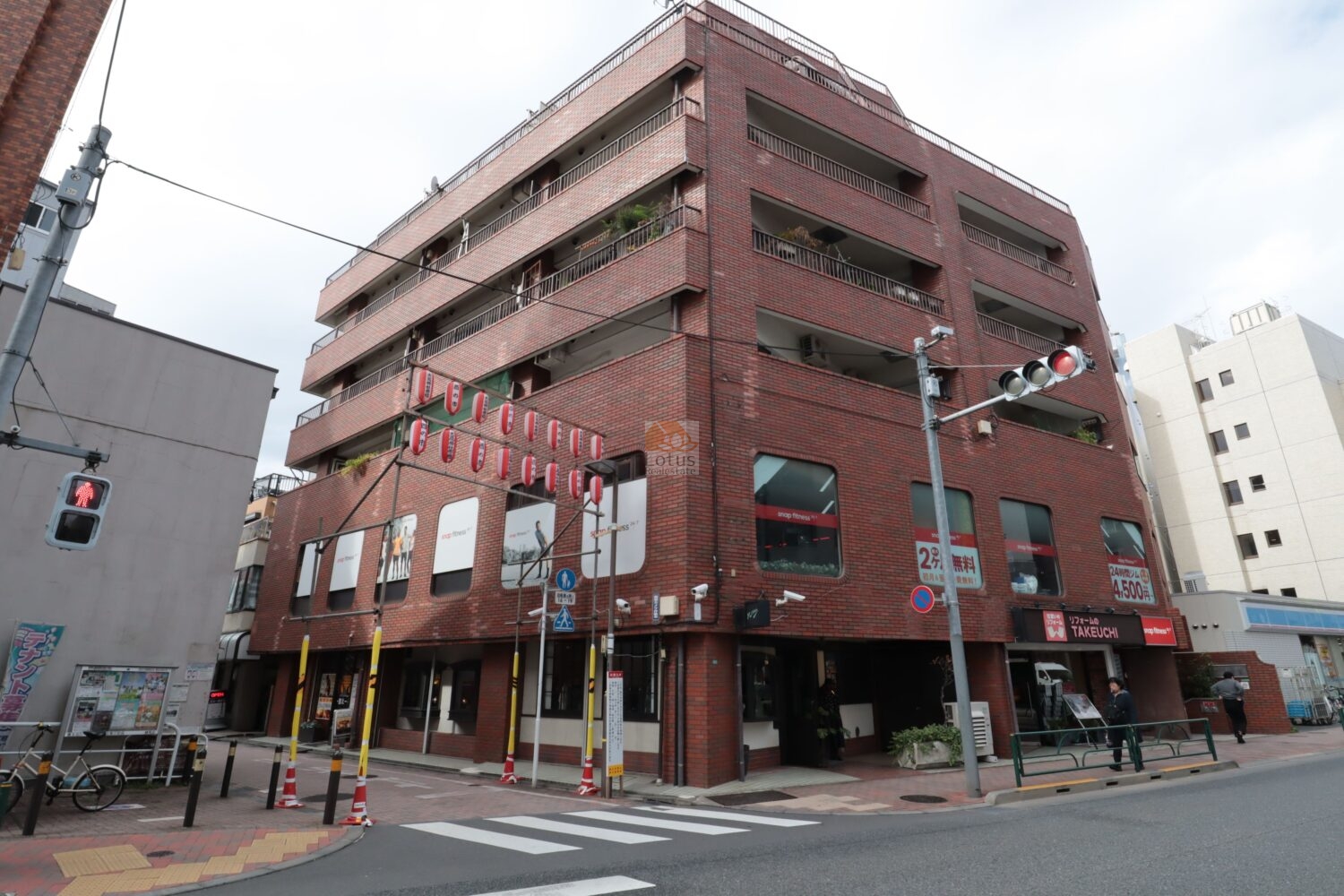 マンション「サンライズ豊玉」の角地立地における外観です。レッドブラウンのタイル仕上げが目立ち、1階テナント・2階以上住戸という構成が視認できます。ファサードの水平に連なるバルコニー開口が建物ボリュームを視覚的に分節しており、街並みに溶け込む印象を受けます。