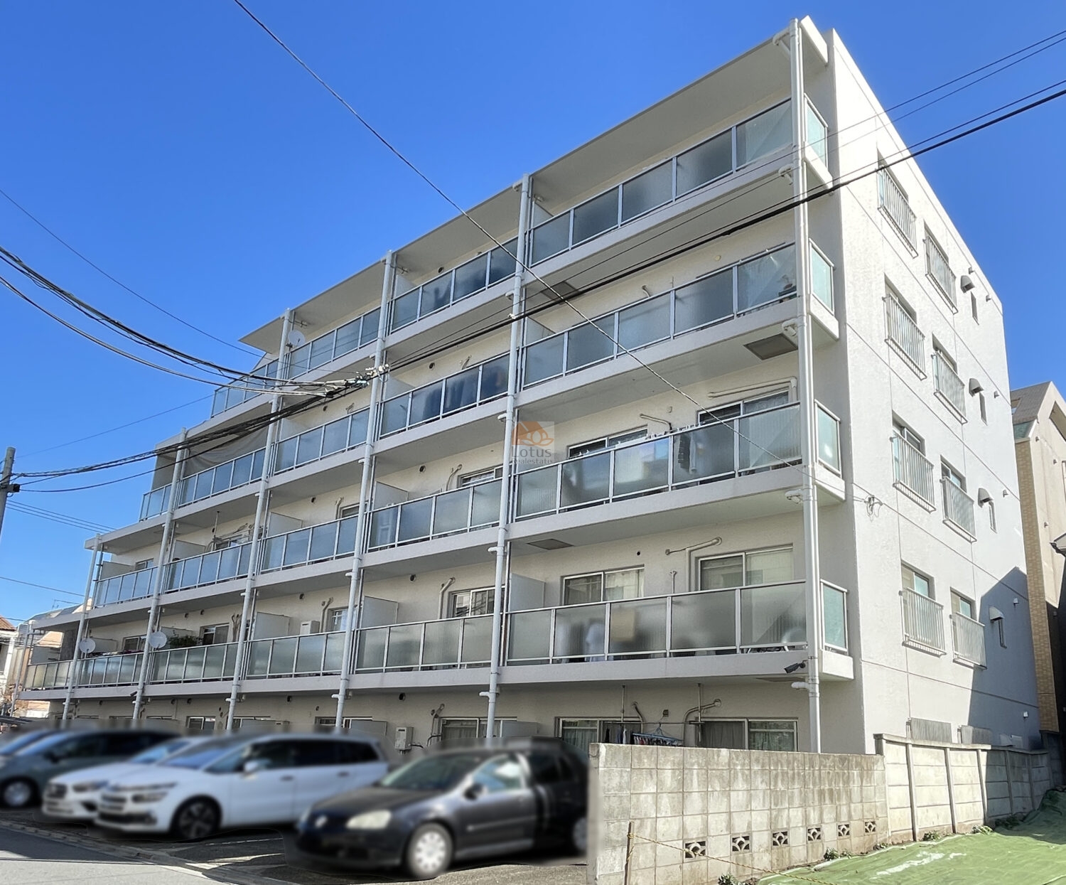 藤和用賀コープⅡの外観。白を基調とした5階建の外観で、ガラス手すりのバルコニーです。建物の前面には駐車スペースが設けられ、複数台の車が停められている様子が見えます。周囲は低層の住宅が混在する落ち着いた街並みで、第一種住居地域らしい穏やかな景観の中に建物が佇んでいます。藤和用賀コープⅡは、昭和56年築、5階建てのマンションです。