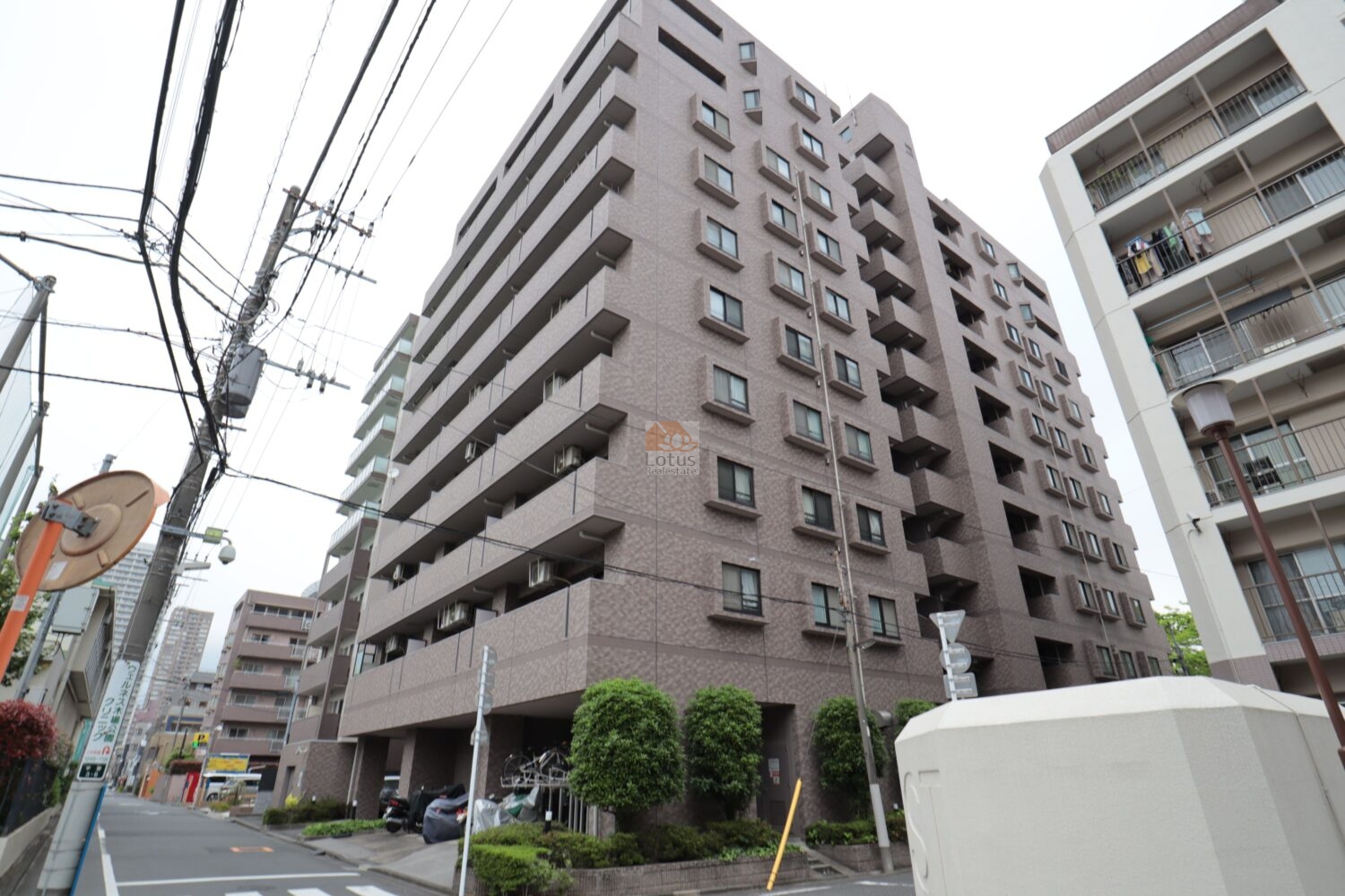 ライオンズマンション木場公園の外観で、ブラウン系タイル貼りの中層建物が交差点角に立地している様子です。バルコニーが水平に連なり、周囲の建物と高さがそろうため街並みに溶け込みます。前面道路は見通しがよく、車両と歩行者の動線が分離された穏やかな印象です。