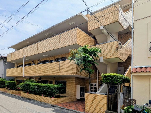 セブンスターハイツ池上の外観。落ち着いたベージュ系タイル貼りの低層建築が街並みに調和し、全戸南向き配置で日当たりにも配慮された造り。植栽帯が建物を囲み、通り沿いにゆとりある空間を確保。セブンスターハイツ池上は、昭和58年築、4階建てのマンションです。