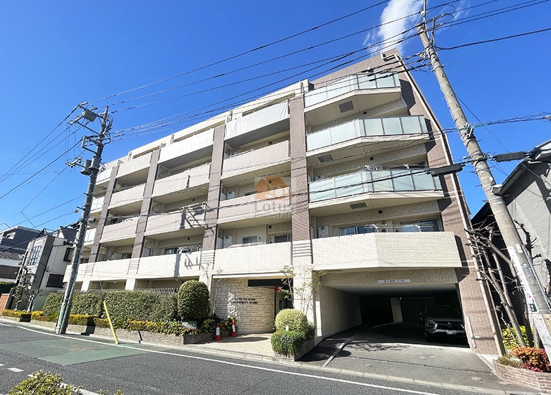 ベージュとブラウンのタイル貼り外観が5層で連なり、右手に車路を兼ねたピロティ開口、歩道側に生垣と低木の植栽、エントランス脇に銘板が見えます。快晴の青空の下、建物全体のボリュームと街区のスケールがそのまま写っています。グローベルザ・シーズン鷺ノ宮パークスは、平成24年築、5階建てのマンションです。