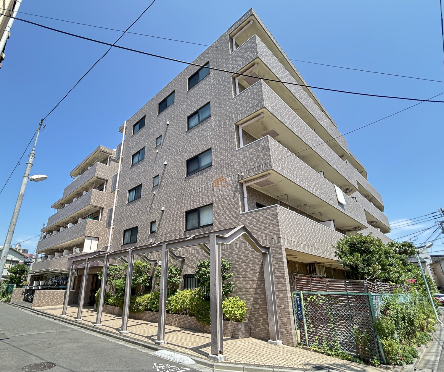 淡いベージュのタイルが整然と貼られた外観で、落ち着いた印象の５階建てマンションです。通り沿いには植栽が続き、足元にはタイル敷きのアプローチが広がっています。南側からの日差しをしっかり受ける角地のため、建物全体が明るく、空の青さが映える雰囲気でした。