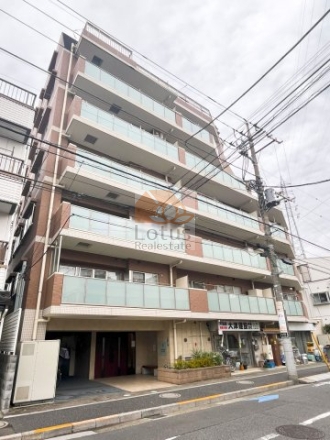 ミオカステーロ西小岩の外観。通り沿いに面した中層の住棟が写っています。正面寄りの外観で、フロストガラスのバルコニーとタイル外壁が確認できます。1階のピロティに出入口があり、歩道沿いに植栽と電柱、上空に電線が通っています。1階にはテナント区画と車路の開口、前面に電柱と電線が見えます。ミオカステーロ西小岩は、平成18年築、7階建てのマンシ
