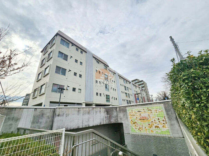 パークハイツ大森の外観。地上4階・地下1階の建物は白を基調とした外観で、シンプルな直線が並ぶ中に小窓がリズムを生んでいます。前面には緑化とタイル壁が設けられ、歩道と住まいを柔らかに区切りながら、都市の生活と居住空間をつなげています。パークハイツ大森は、昭和54年築、4階建て（地下1階付き）のマンションです。