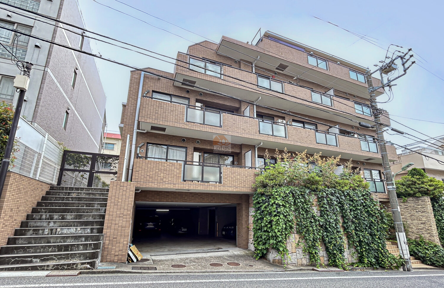 東急ドエル・アルス久が原カリエールの外観。緩やかな坂道に面して建つ外観は、茶系のタイル貼りが整然と並び、バルコニーにはガラス手すりが採用されています。足元には緑が配され、石造りの擁壁を覆うツタが、街並みに落ち着いた雰囲気を添えています。東急ドエル・アルス久が原カリエールは、平成11年築、5階建てのマンションです。