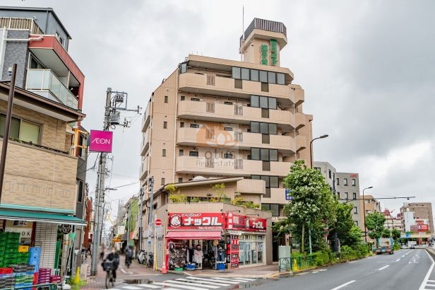 環七通り沿いに建つ建物全景で、ベージュ系タイル貼りの外壁とバルコニーの段差構成が特徴的です。低層部には作業服店「ナックル」が入居しており、周囲の店舗や通りの風景と一体となった街並みを形成しています。朝日プラザ旗の台は、昭和61年築、7階建てのマンションです。