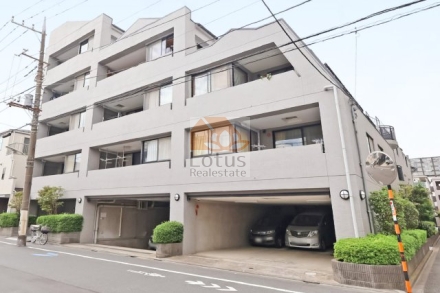 通り沿いに明るいグレーの外壁が続き、1階は奥行きのあるピロティ駐車場になっています。開口の手前に円形ミラーと植栽のプランター、歩道の矢印表示と白線が見えます。上階はバルコニーの連なりとサッシが整列し、電柱と配線が建物前を横切ります。マイキャッスル池上ガーデンコートは、平成13年築、6階建てのマンションです。