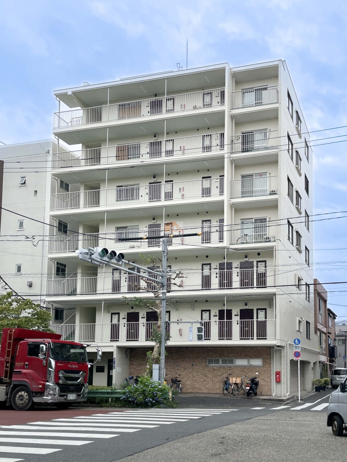 白いタイル調の外壁が印象的な中層マンションです。建物は地上6階建てで、バルコニーと外廊下が整然と並んでいます。1階部分にはレンガ調の外壁が施され、駐輪スペースが設けられています。前面道路には横断歩道と信号機があり、交通量のある通りに面していますが、歩道も整備され、安全に配慮された環境が見て取れます。セントラル王子は、昭和59年築、7階建てのマンションです。