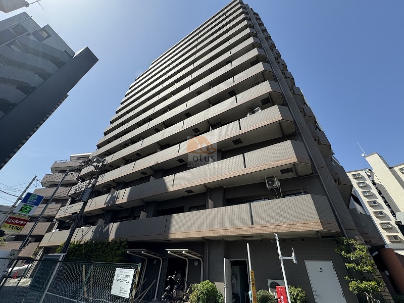 淡いグレー系タイルが整然と積まれた外観で、直線的なバルコニーの段差が光の角度によって陰影を作る雰囲気です。京急本線「大森町」駅徒歩1分という立地らしく、視界には駅前の商業動線が入りやすく、暮らしの導線がわかりやすいと感じました。中層～高層帯の住宅が続くエリアとの距離感は、都心寄りの生活動線を意識しやすい印象です。マイキャッスル大森町エクセレントステージは、平成14年築、14階建てのマンションです。